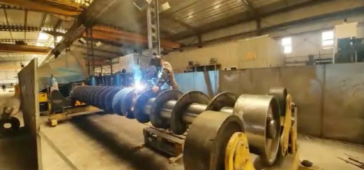 A welder working on a large screw press shaft in a workshop